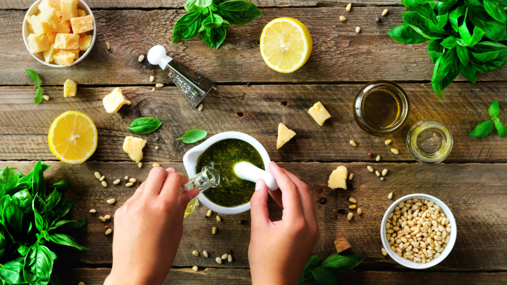 Woman hands making italian pesto in bowl. Ingredients - basil, lemon, parmesan, pine nuts, garlic, olive oil and salt on rustic wooden background. Top view, flat lay, copyspace